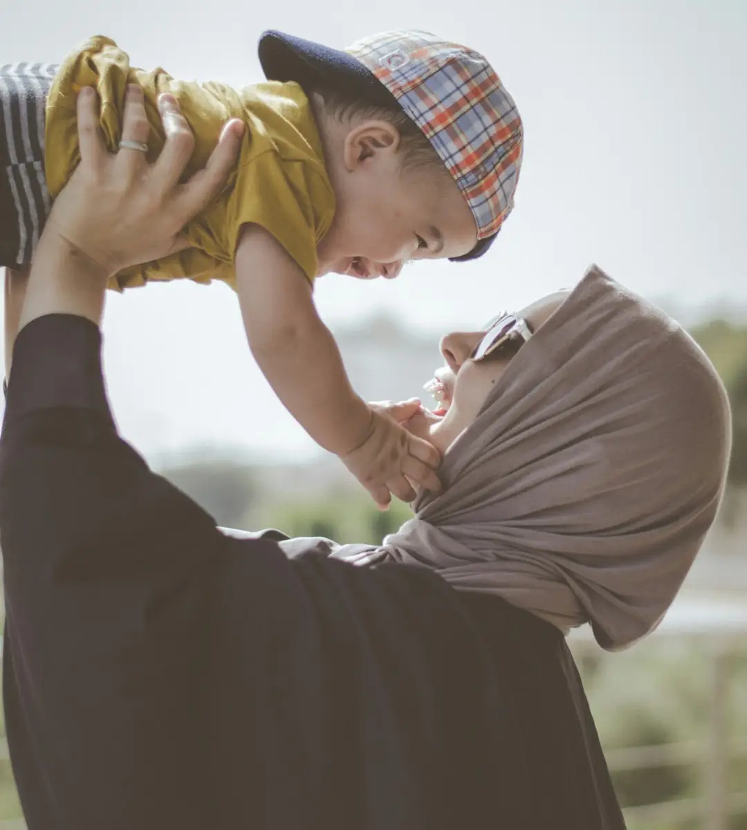 Woman wearing hijab and sunglasses holding baby in the air, both smiling