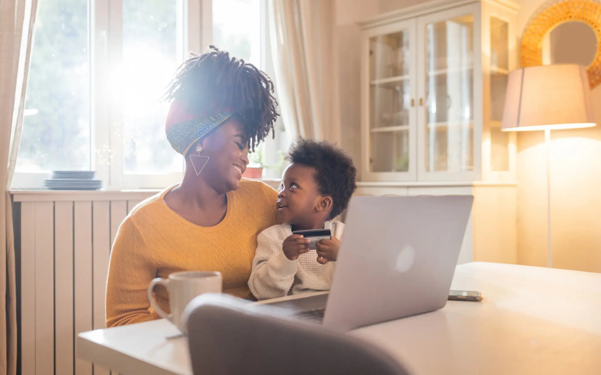 Smiling woman with baby on her lap holding a credit card in front of a laptop on the table