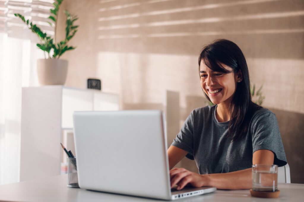 Woman smiling during a virtual coaching session, seated at a laptop in a calm home office environment.