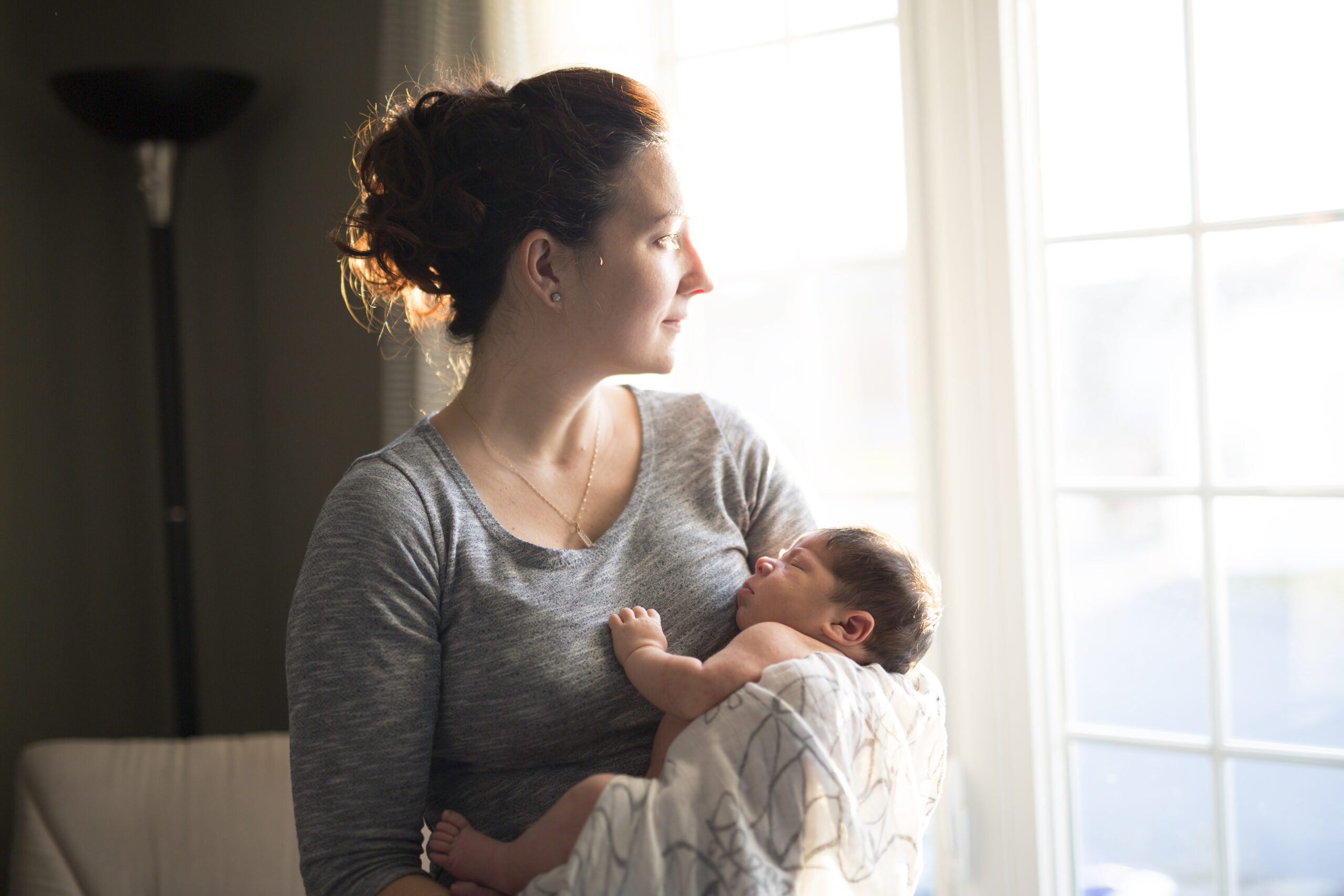 Mother holding baby while gazing out a window, reflecting emotional overwhelm and identity shifts in early motherhood