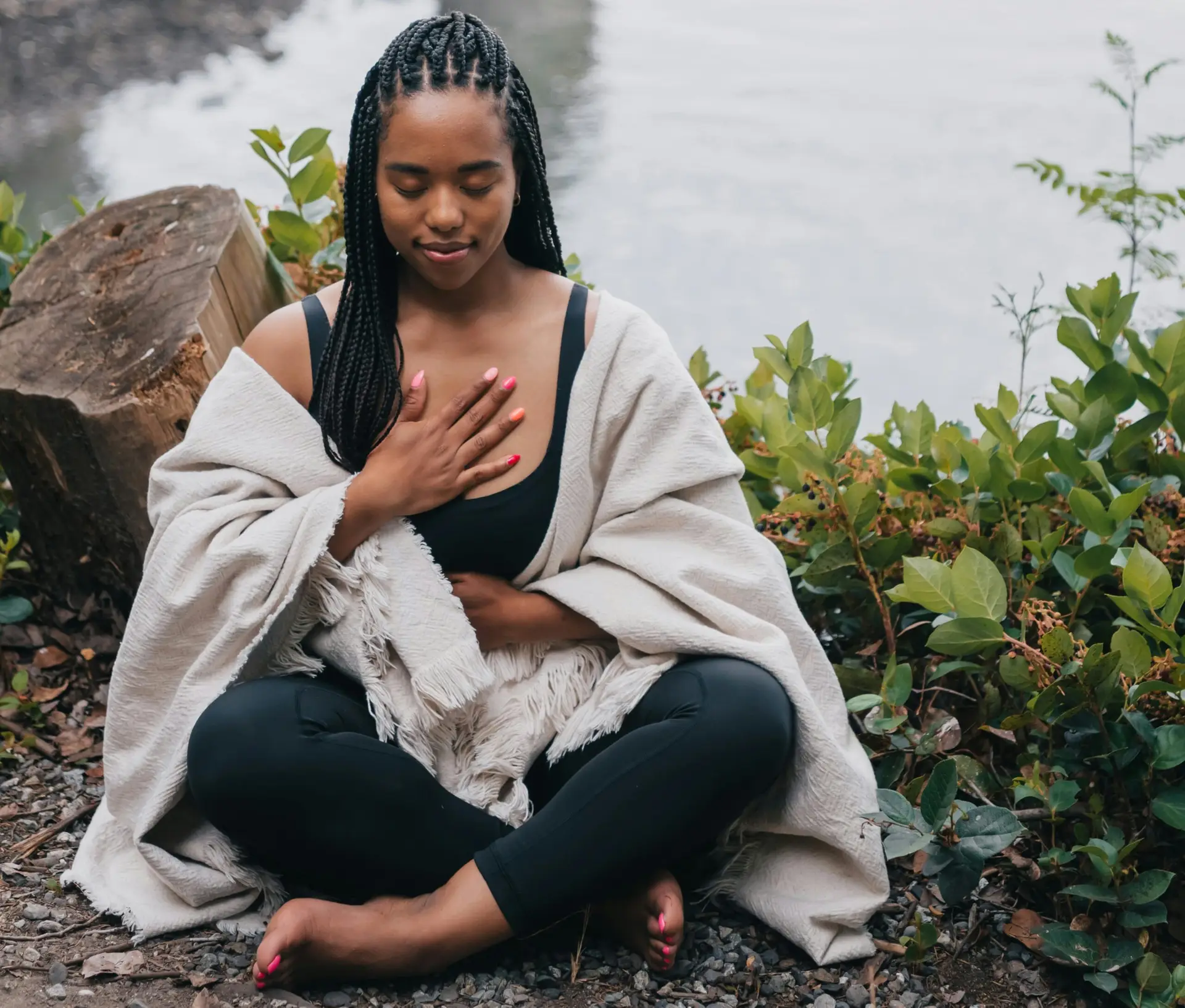 Young Black woman sitting cross legged on ground with eyes closed, one hand on heart, one hand on stomach, and serene