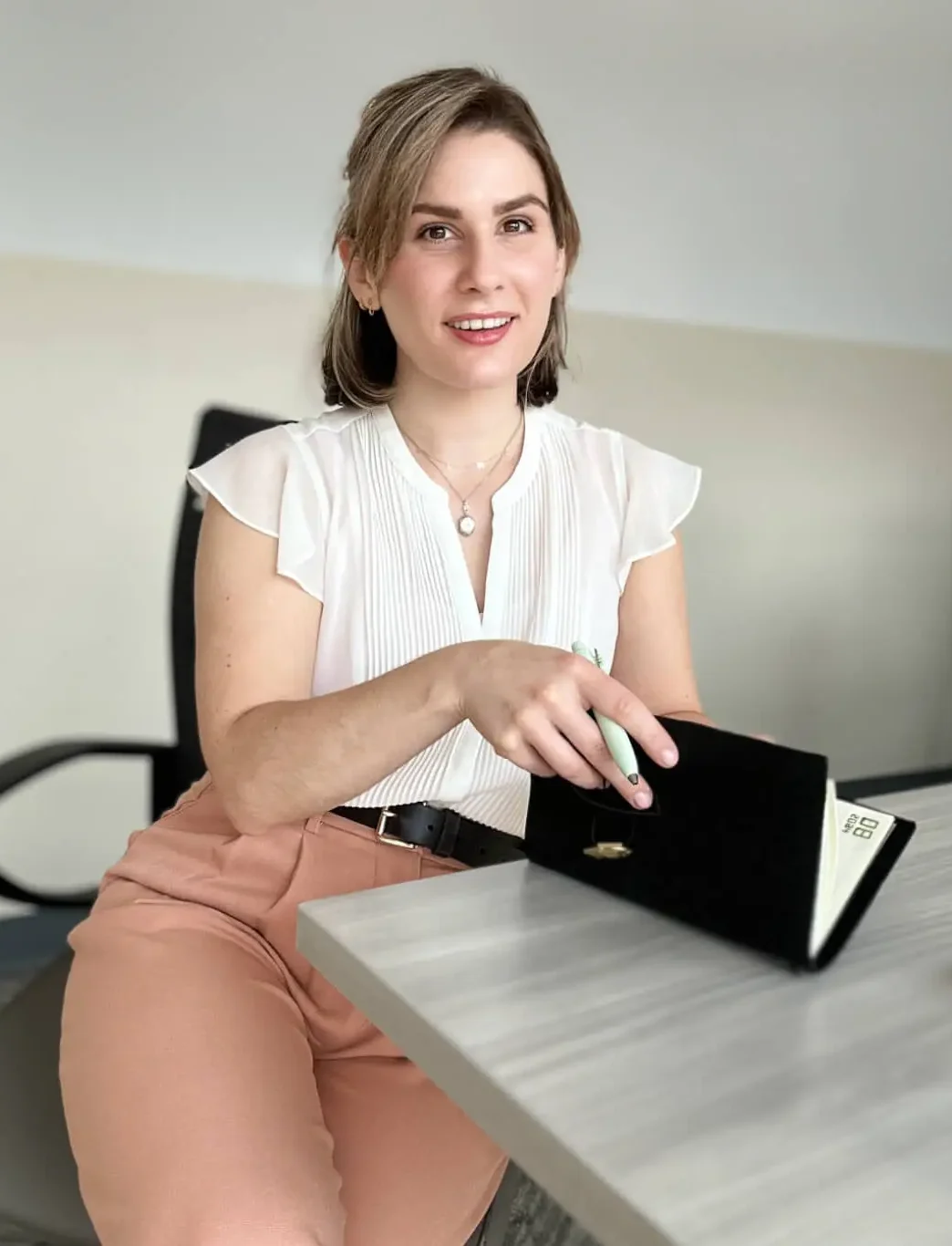 Young woman smiling and sitting at desk with pen in hand and opening a journal while looking at viewer