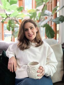 A woman with brown hair and a warm smile sits on a cozy couch holding a white owl-shaped mug, surrounded by soft textures and leafy indoor plants in natural light.