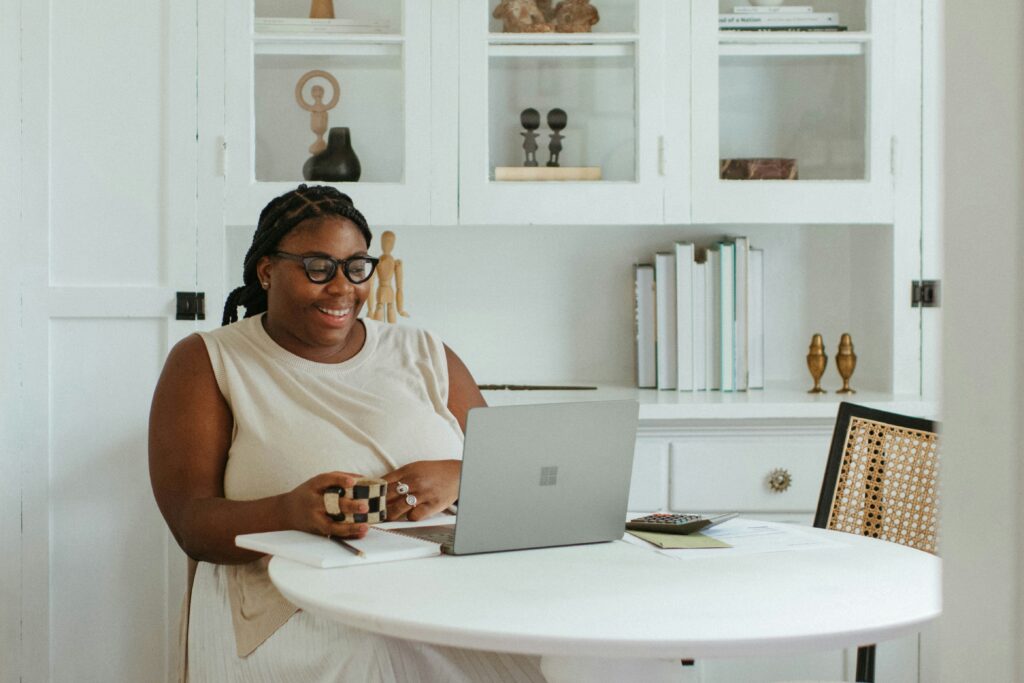 A woman sitting at a round table during a virtual therapy session, using a laptop in a bright and calming home office.